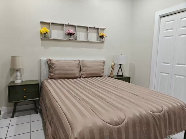Quarto com cama, mesas de cabeceira, cômoda, prateleira com flores e porta.