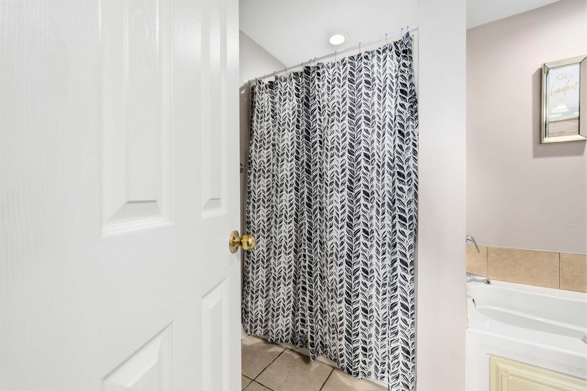 A bathroom with a white door, a shower curtain with a leaf pattern, and a framed picture on the wall.