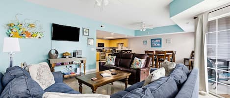 A living room with a sofa, coffee table, and a TV. There is a kitchen in the background.