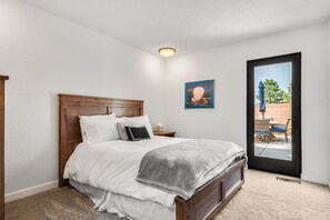 A bedroom with a wooden bed, white bedding, a gray throw blanket, a nightstand, a framed picture, and a door leading to an outdoor area. - Sandia Vista Retreat 5 BDR 4.5 Bathroom, Hot Tub, Game Room (Albuquerque)