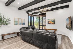 A modern living room with a sectional sofa, a ceiling fan, large windows, and artwork on the walls.