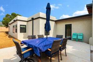 An outdoor patio with a table set for four, surrounded by wicker chairs, a blue umbrella, and a small building with a door and windows.