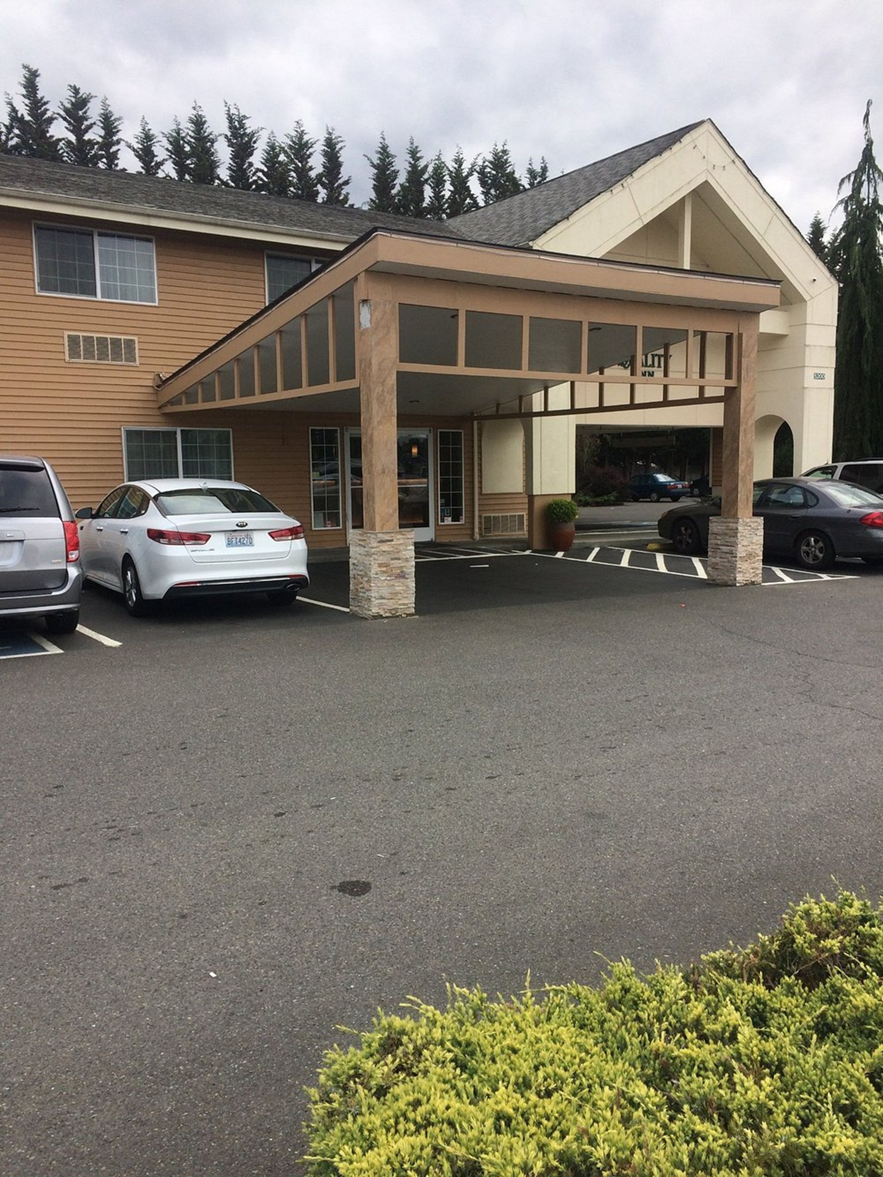 A hotel entrance with a covered parking area, a building with a beige facade, and a row of parked cars.