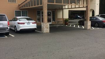 A hotel entrance with a covered parking area, a building with a beige facade, and a row of parked cars.