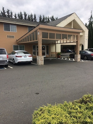 A hotel entrance with a covered parking area, a building with a beige facade, and a row of parked cars.
