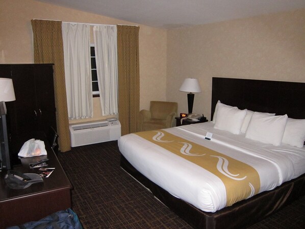 A hotel room with a bed, a desk, a chair, a nightstand with a lamp, and a window with curtains.