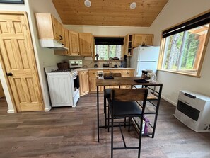 Cocina compacta con armarios de madera, estufa, microondas y refrigerador. Hay una mesa de comedor con sillas y un ventanal con vistas al exterior.