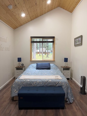 Un dormitorio con una cama grande, una ventana con vistas a árboles, dos mesitas de noche con lámparas y una pared con una cita.