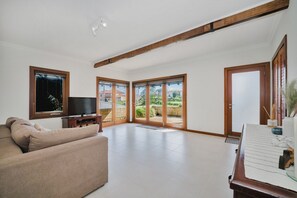 A spacious living room with a sofa, a television, and a piano. There are large sliding glass doors leading to a balcony with a view of houses. - Ocean-View 3-Bed House in Halls Head (Halls Head)