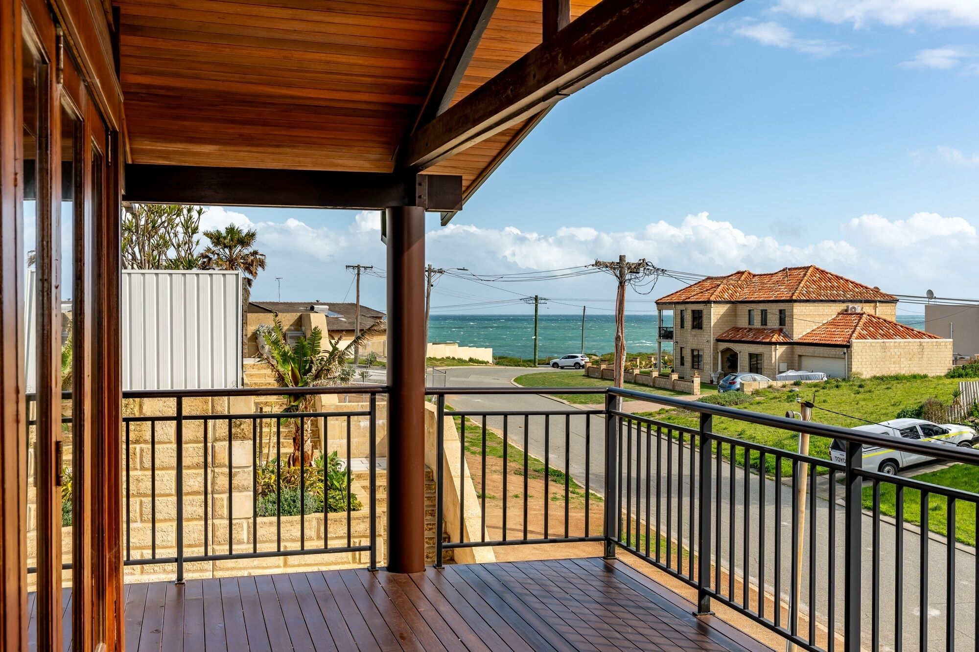 A wooden balcony with a view of a coastal road and a house with a tiled roof.