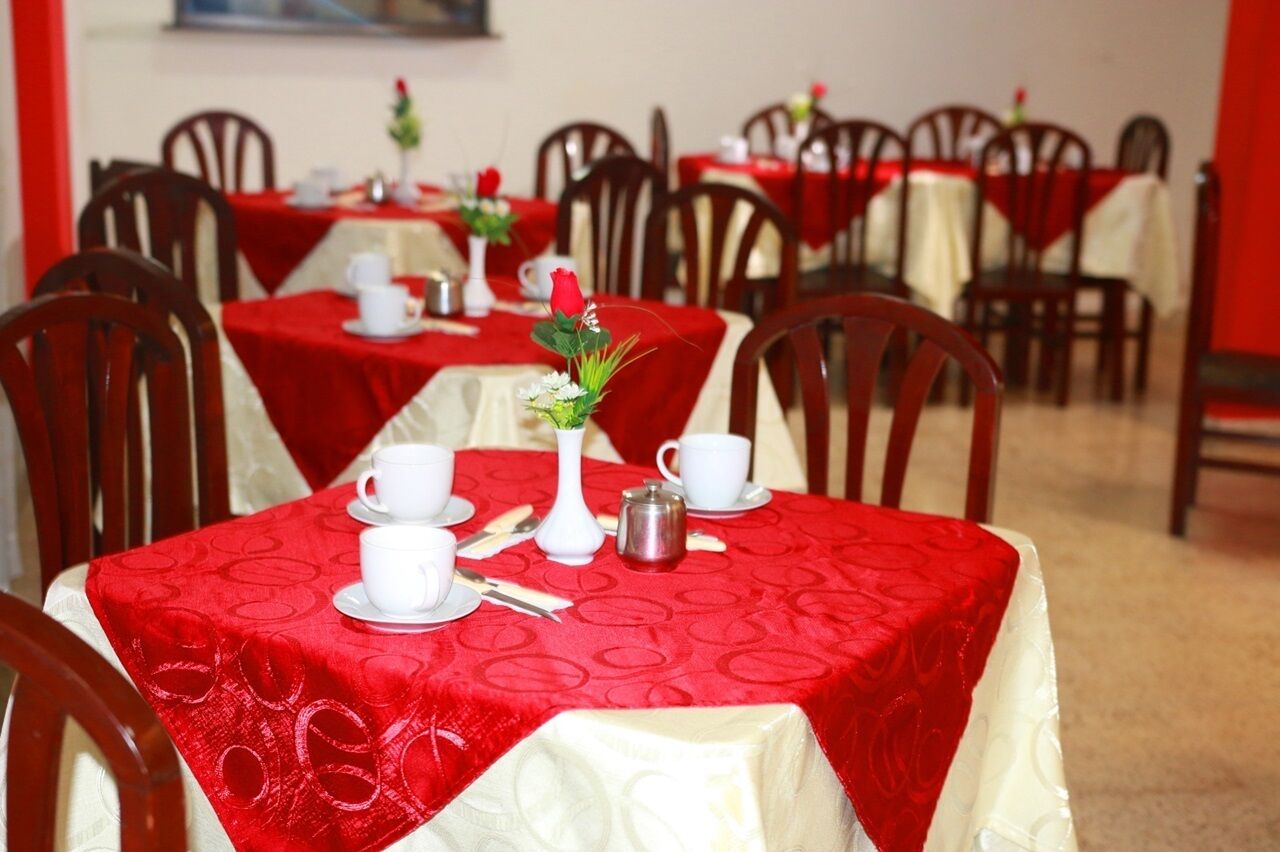Un comedor con mesas dispuestas para dos, cada una con tazas, platillos y un pequeño jarrón con una rosa roja.