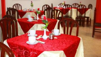 Un comedor con mesas dispuestas para dos, cada una con tazas, platillos y un pequeño jarrón con una rosa roja.