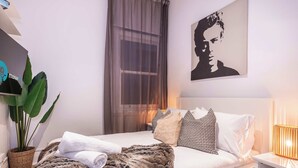 A modern bedroom with a bed, pillows, a nightstand, a lamp, a large window with curtains, and a wall-mounted artwork. - Quiet Art Deco 2BR Retreat in the Heart of Potts Point (Potts Point)