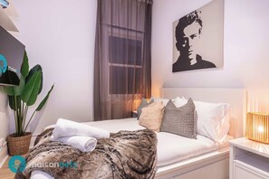 Ett modernt sovrum med en säng, kuddar, ett sängbord, en lampa, ett stort fönster med gardiner och ett konstverk som hänger på väggen.