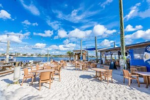En café ved stranden med træborde og stole, havudsigt og en blå himmel med skyer.