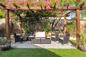 A covered patio with wicker furniture, a stone wall, and flowering plants. - 4BR Family Getaway  Private Office, Playground & Backyard (Thousand Oaks)
