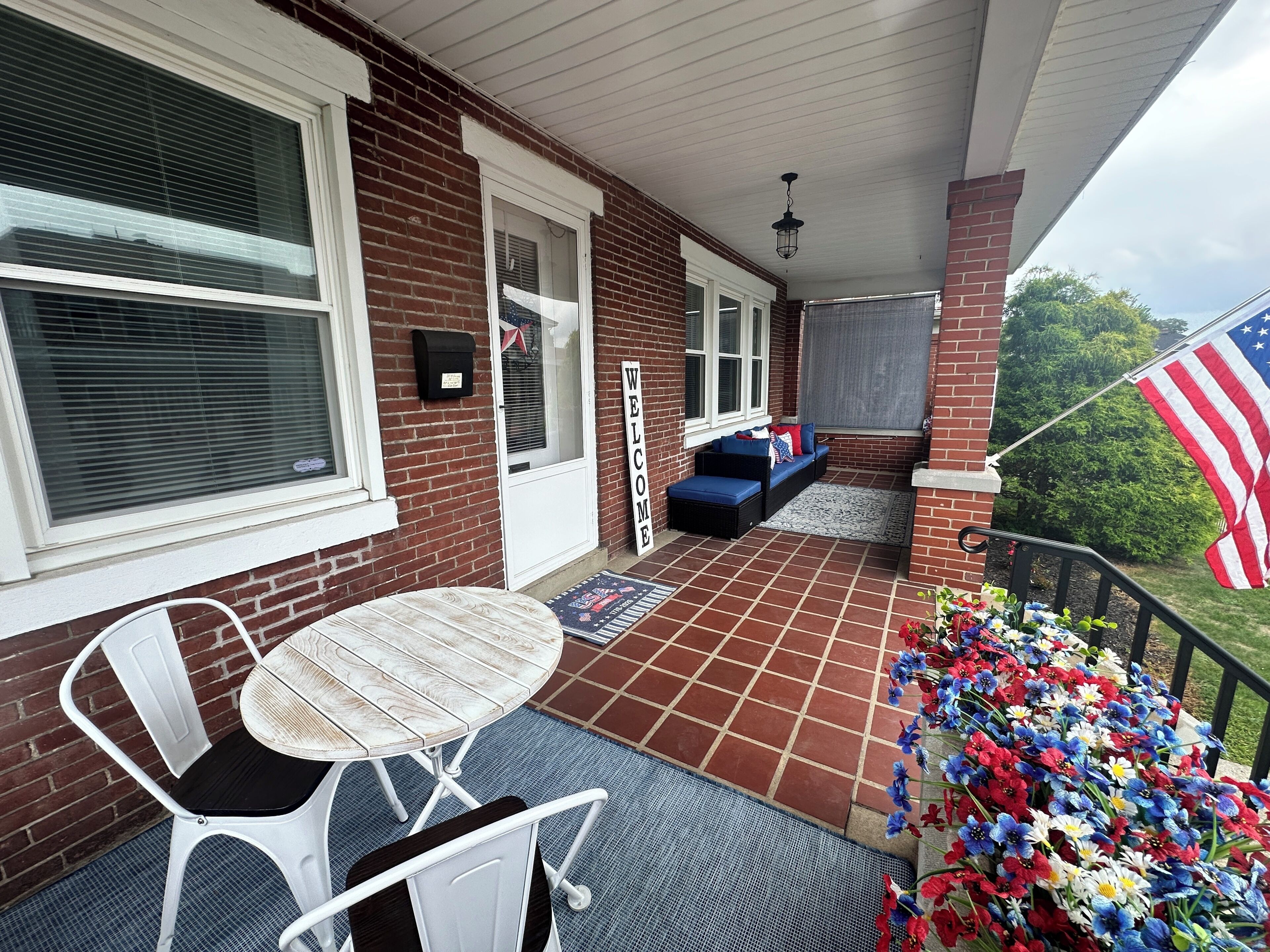 A covered patio with a brick wall, a white metal table and chair, a blue sofa, and an American flag.