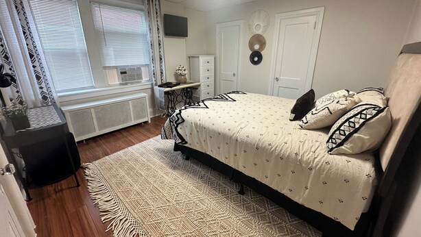 A bedroom with a bed, bedside table, dresser, radiator, and a window with curtains.