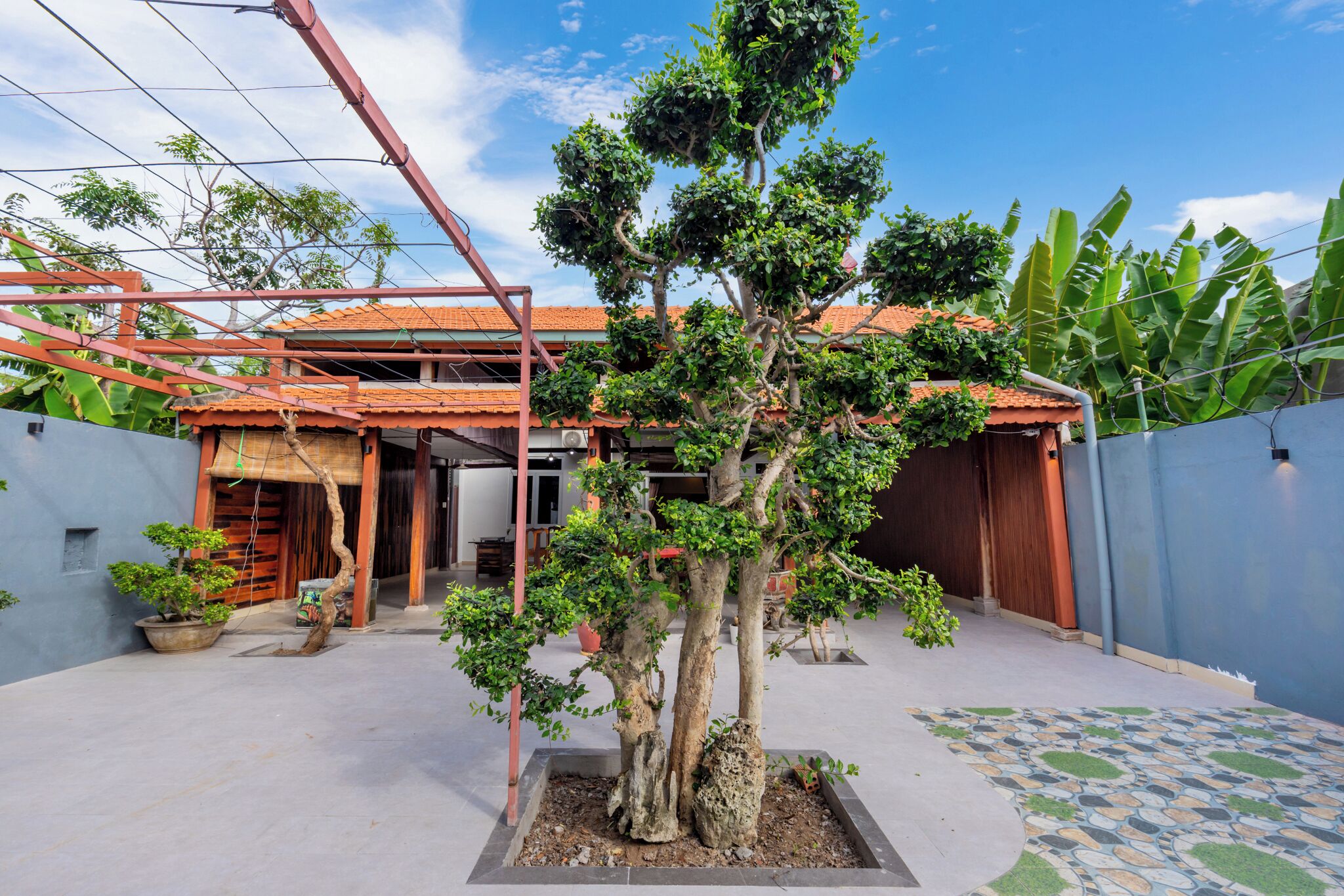 A residential property with a covered patio area, a house with a tiled roof, and a garden with a central tree.
