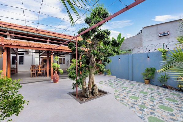 A covered patio area with a dining table and chairs, surrounded by potted plants and a tree.
