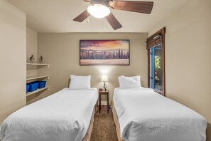 A hotel room with two beds, a ceiling fan, a painting of a snowy landscape, a nightstand with a lamp, and a window with a view of greenery.