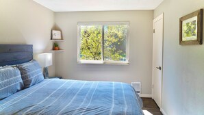 Une chambre à coucher comprenant un lit, une table de chevet, une lampe, une fenêtre donnant sur des arbres et une porte.