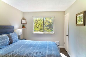 Ein Schlafzimmer mit einem Bett, einem Nachttisch, einer Lampe, einem Fenster mit Blick auf Bäume und einer Tür.