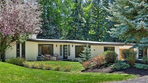 A modern two-story house with a covered patio, surrounded by a well-maintained garden and tall evergreen trees. - Hulen Hideaway | Hot Tub | Perfect Location (Ketchum)