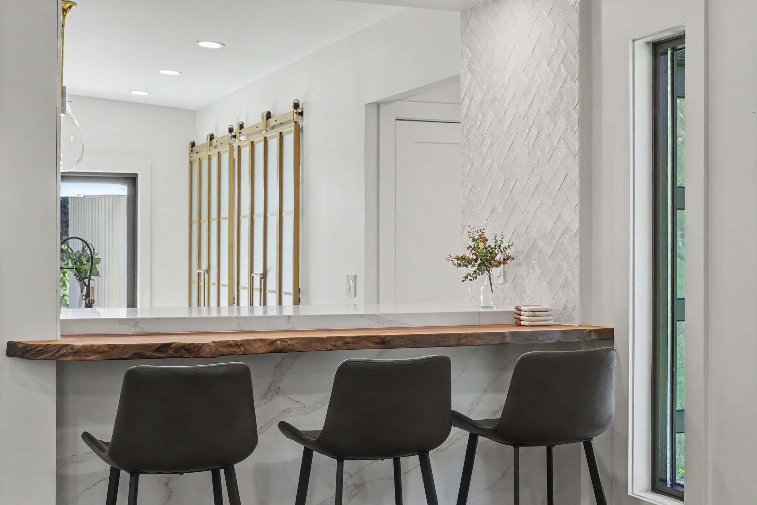 A modern interior with a wooden counter, three black chairs, and a large mirror reflecting the room.
