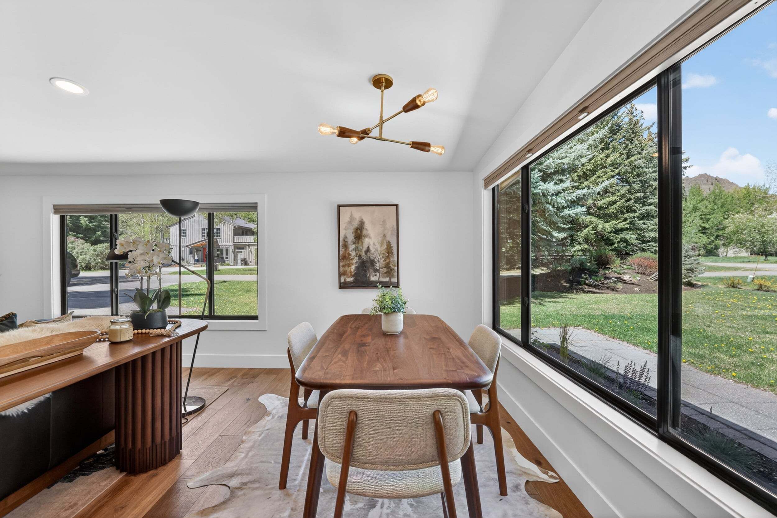 Un coin repas avec une table en bois, des chaises, une suspension et une grande fenêtre donnant sur un jardin et des bâtiments.