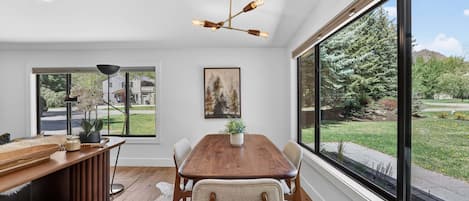 Un comedor con una mesa de madera, sillas, una lámpara colgante y un ventanal con vistas a un jardín y edificios.