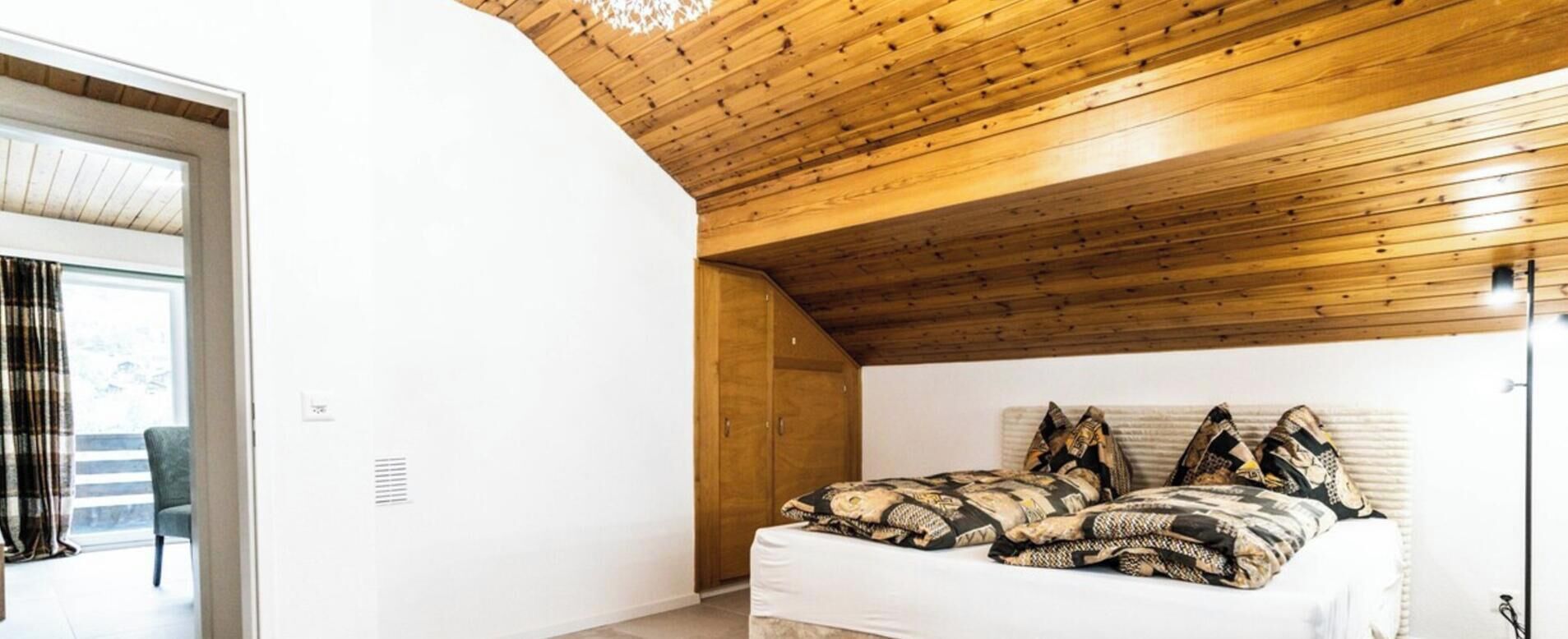 Une chambre avec un plafond en bois incliné, un lit avec une literie à motifs et une vue sur l’extérieur par une porte ouverte.