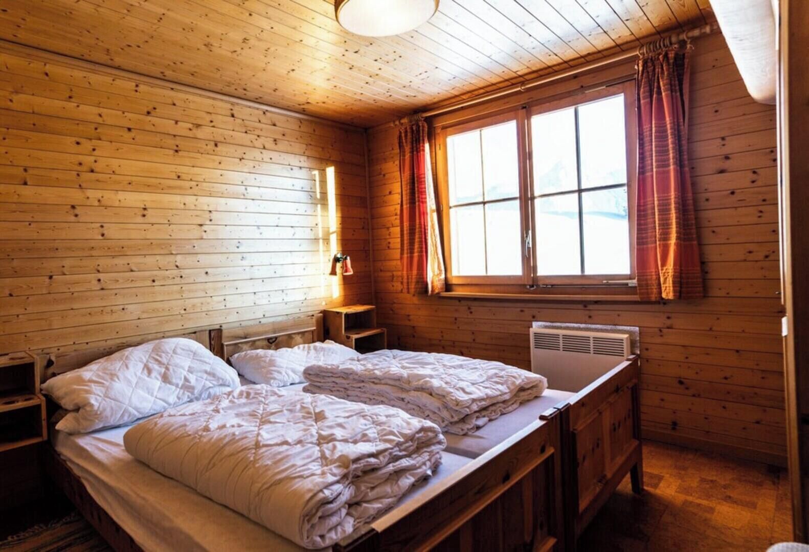 Un dormitorio de cabaña de madera con cama, mesita de noche, ventana con cortinas y un calentador fijado a la pared.