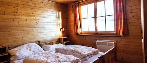 Un dormitorio de cabaña de madera con cama, mesita de noche, ventana con cortinas y un calentador fijado a la pared.