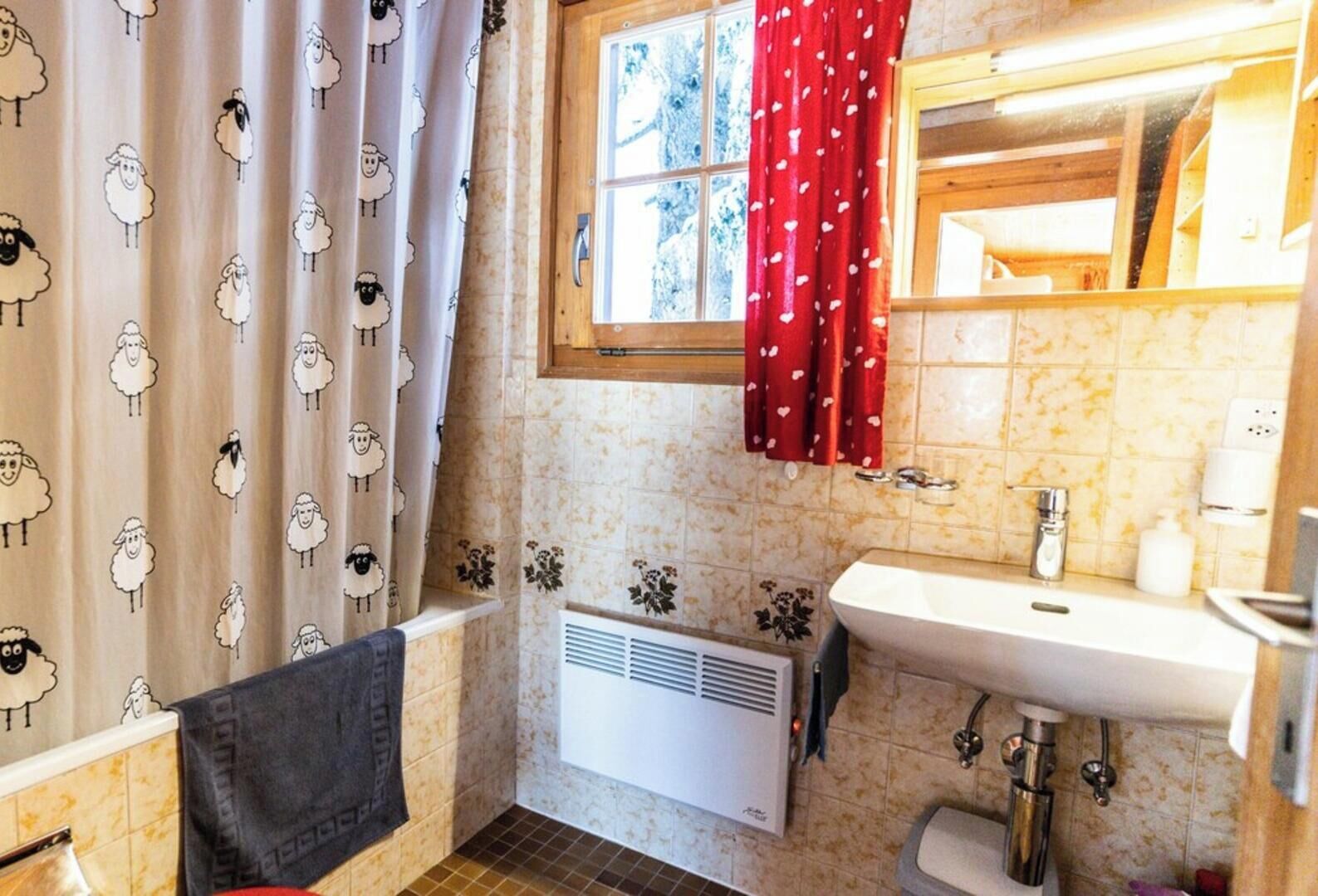 Un baño con cortina de ducha estampada con motivos de ovejas, una cortina roja con corazones blancos, una ventana que da a zonas verdes, un radiador blanco y un lavabo con espejo encima.