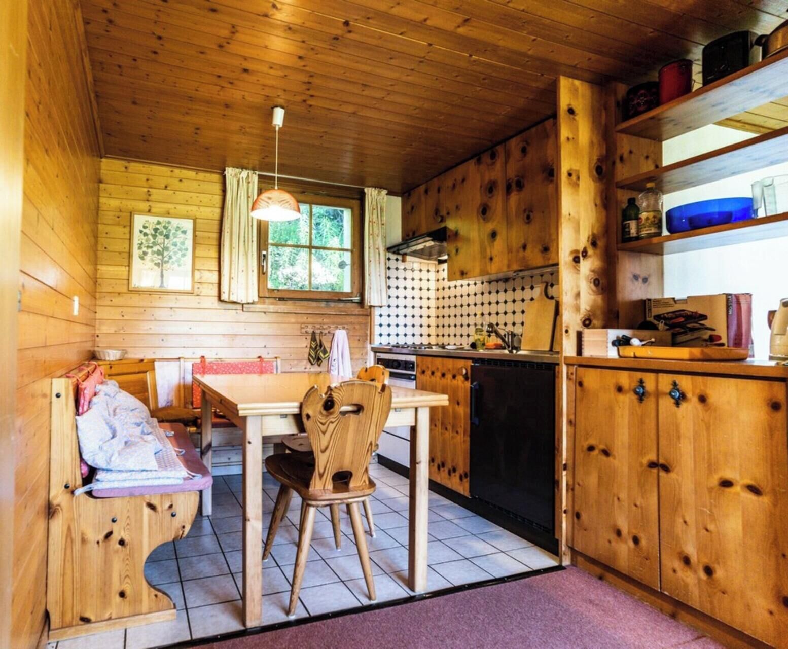 A wooden cabin interior with a dining table, chairs, a kitchen area, and a window with curtains.