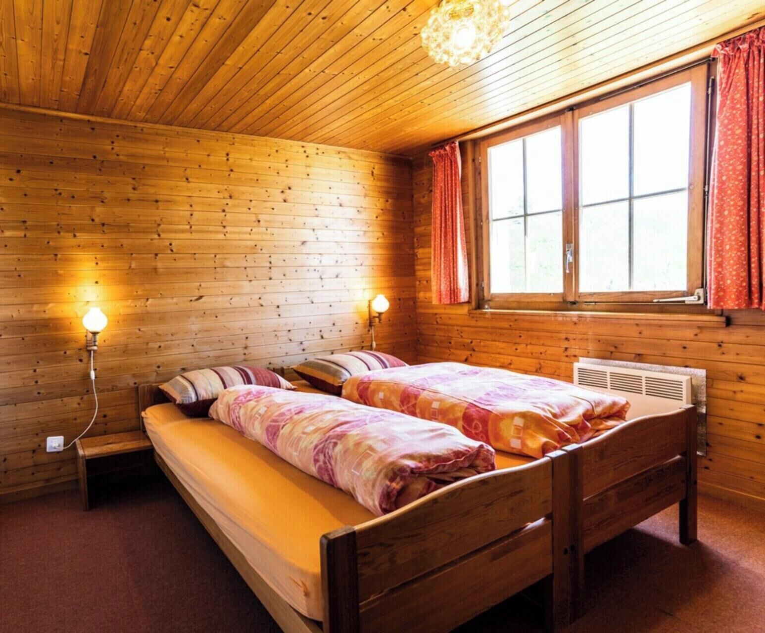 A wooden room with two beds, a window with curtains, and wall-mounted lamps.