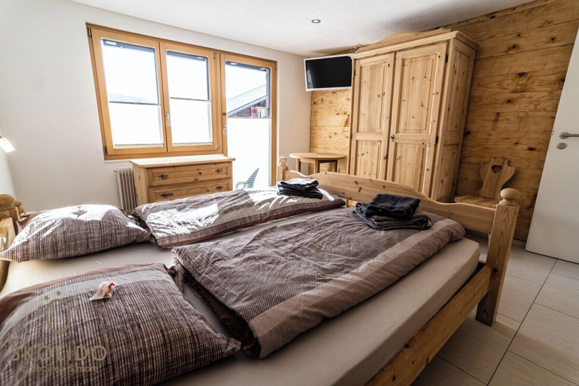 A bedroom with a wooden bed, a TV mounted on the wall, and a window with blinds.
