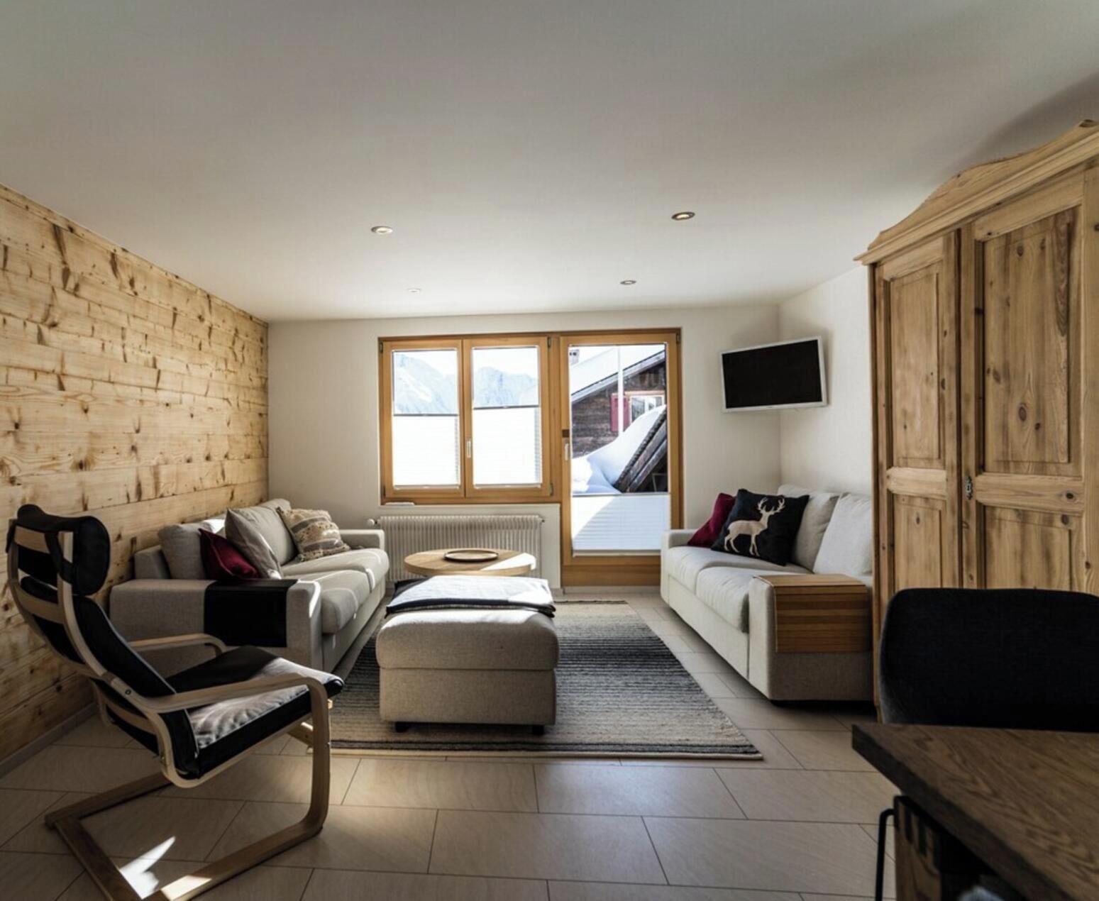 Ein modernes Wohnzimmer mit einer Holzwand, einem Sofa, einem Sessel, einem Fernseher und Blick auf eine Skiseite.