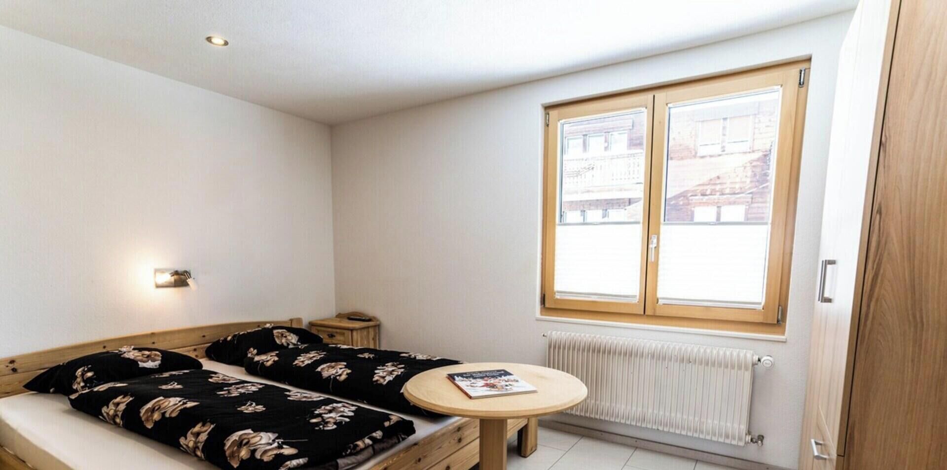 A small, tidy room with a bed, a bedside table, a round table, and a window with a view of buildings.