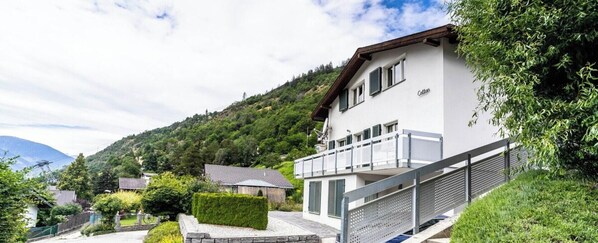 A modern two-story house with a balcony, surrounded by lush greenery and a mountainous backdrop. - Alpine Comfort & Style, Wallis 1053 (Wallis)