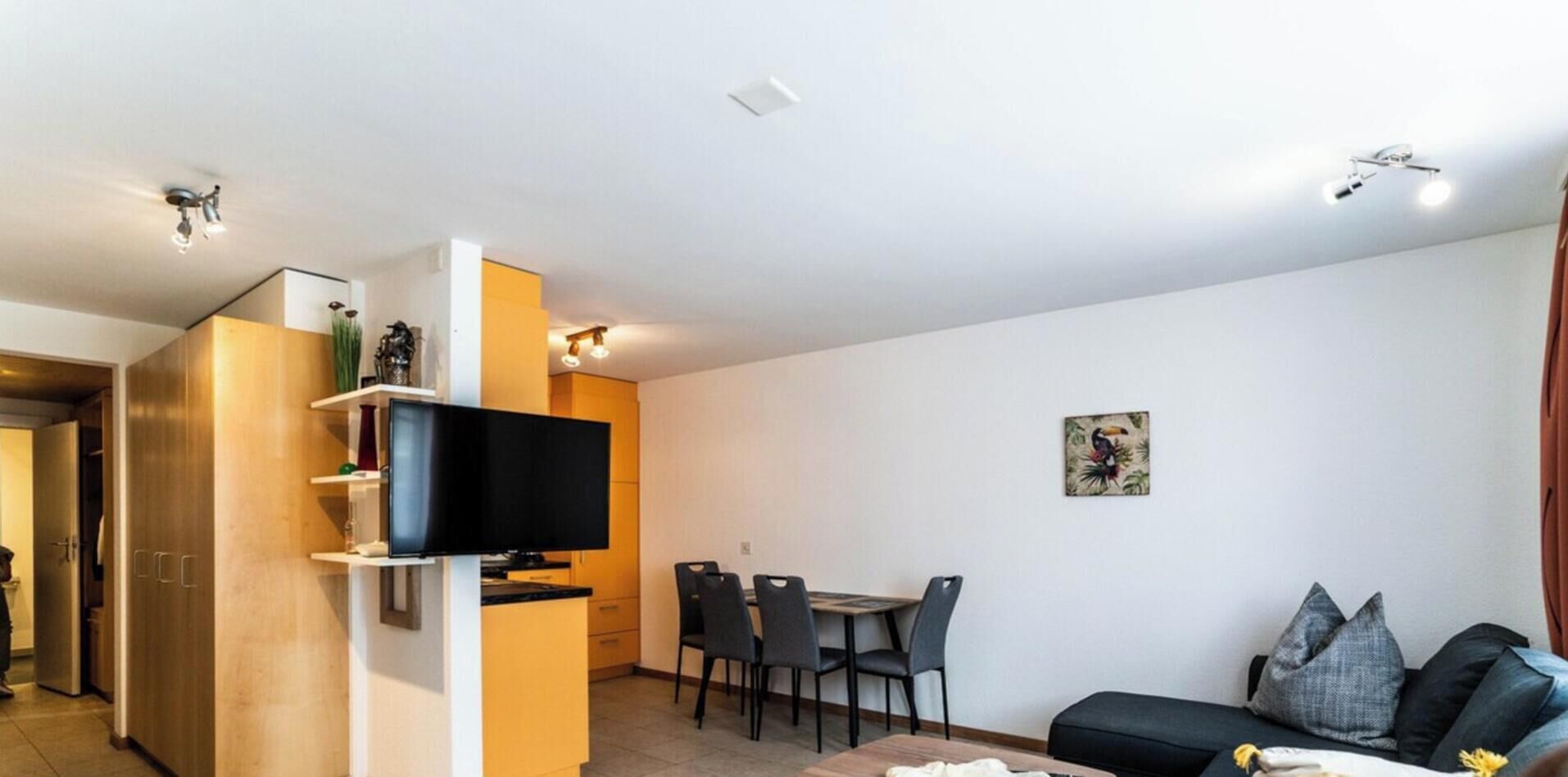 Un salon moderne avec un téléviseur à écran plat fixé sur un meuble en bois, un coin repas avec une table et des chaises, et un canapé avec des coussins.