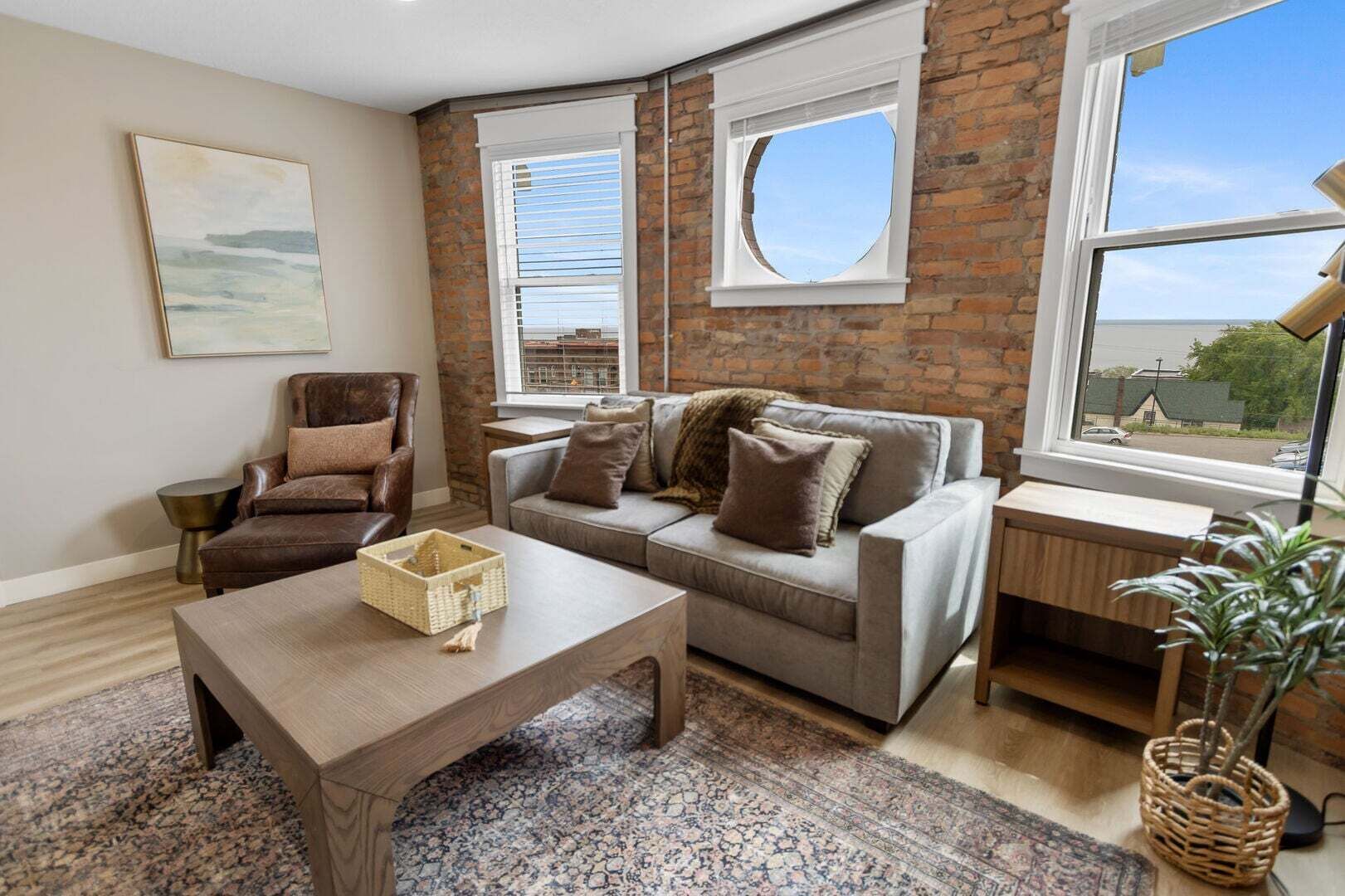 A living room with a brick wall, a grey sofa, a brown leather chair, a wooden coffee table, and a view through a window.
