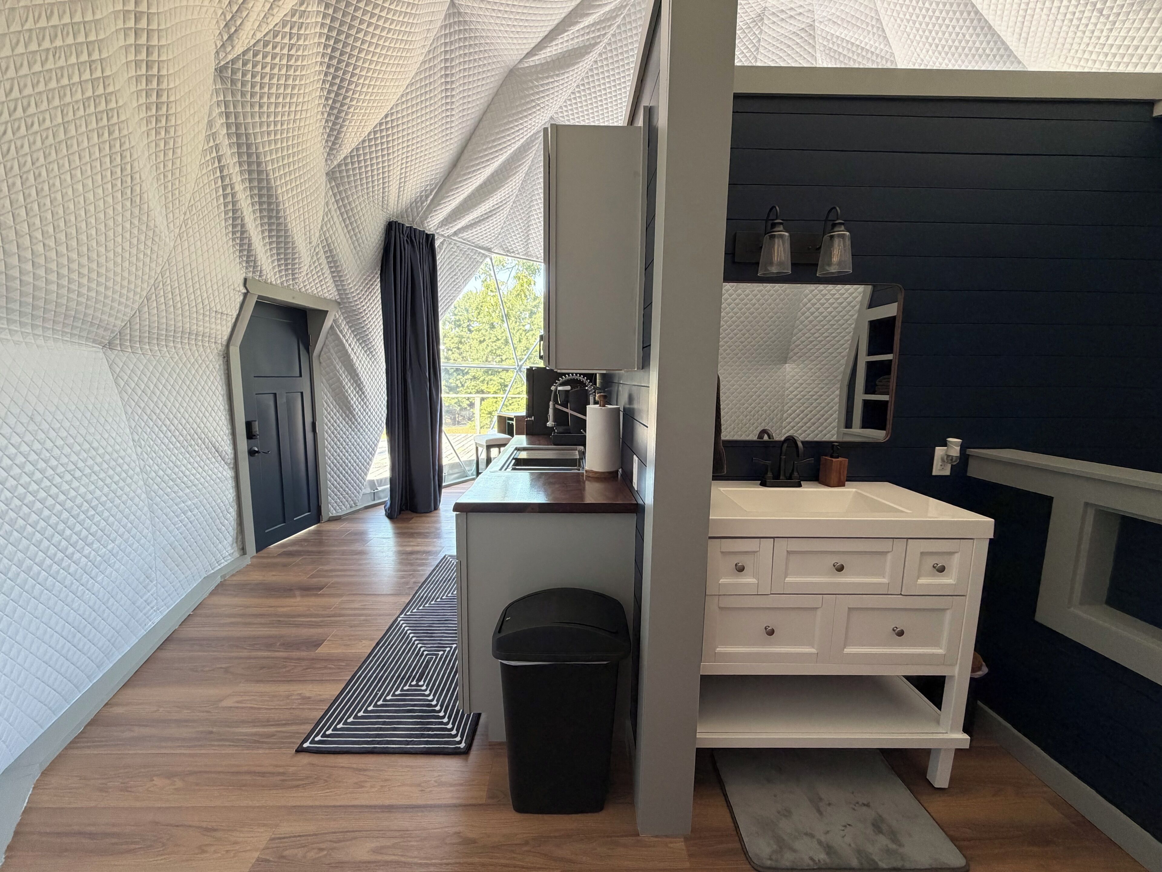 A modern bathroom with a white vanity, a mirror, and a sink. There is a black trash bin and a patterned rug on the floor.