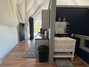 A modern bathroom with a white vanity, a mirror, and a sink. There is a black trash bin and a patterned rug on the floor.