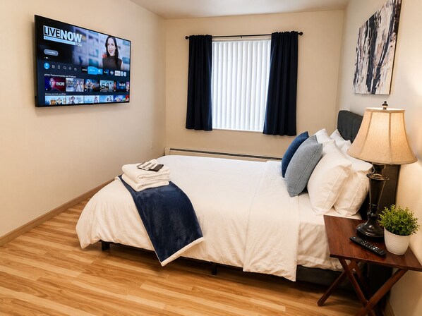 A hotel room with a bed, a TV mounted on the wall displaying a streaming service, a window with curtains, and a bedside table with a lamp and remote control.