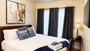 A hotel room with a bed, a bedside table with a lamp, a painting on the wall, and a window with curtains.