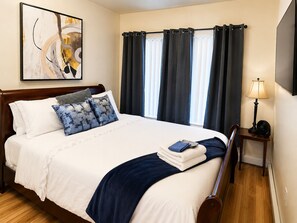 A hotel room with a bed, a bedside table with a lamp, a painting on the wall, and a window with curtains.