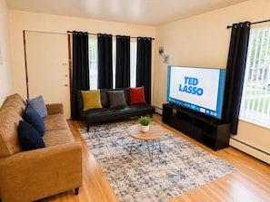 A living room with a sofa, a coffee table, a TV displaying 'TED LASSO,' and a window with curtains.