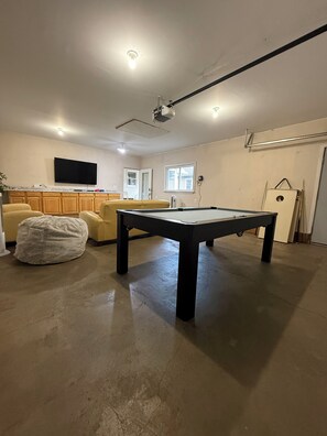 Ein Zimmer mit Billardtisch, einer Couch, einem Fernseher und einem Deckenprojektor.